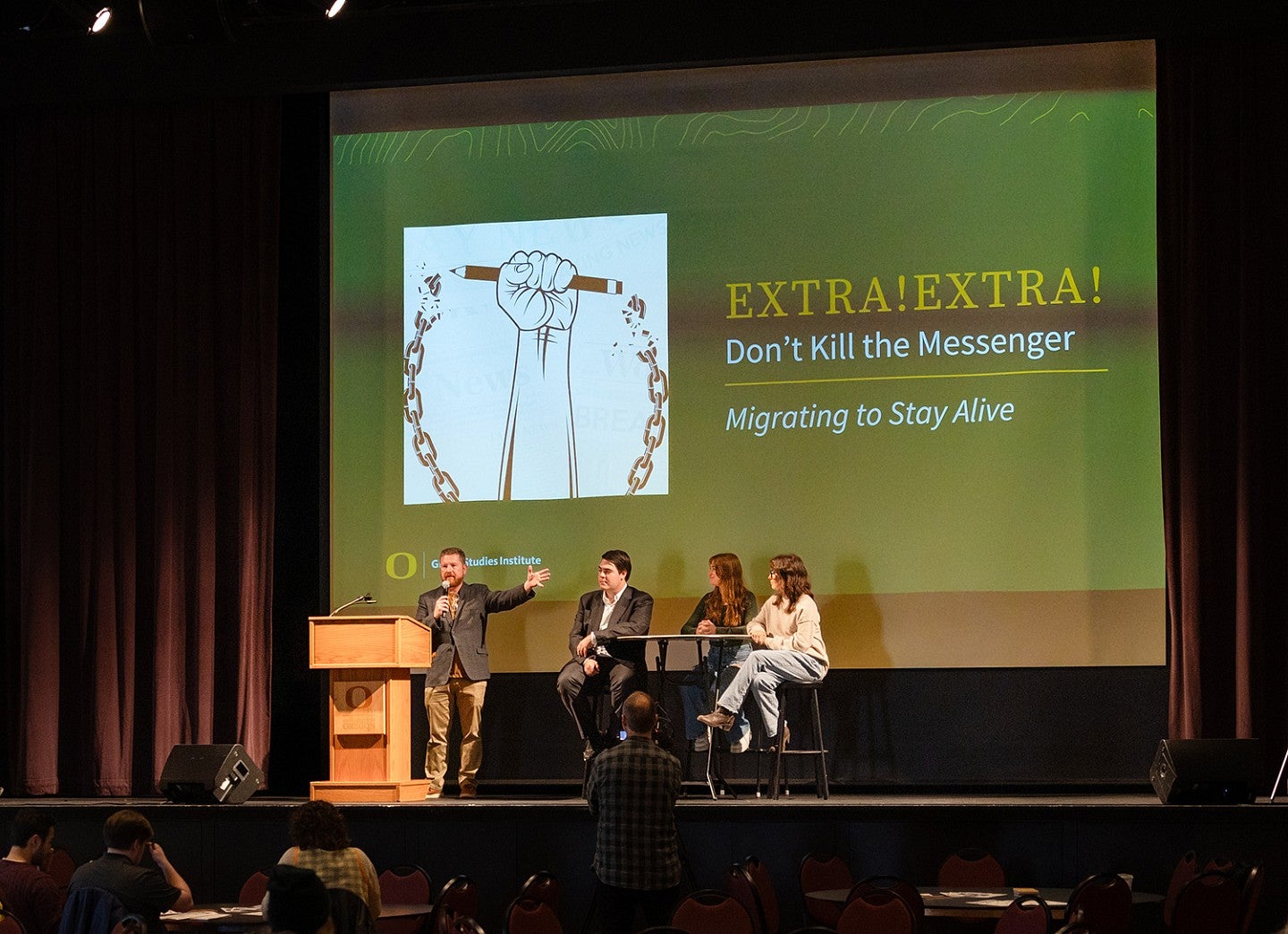 Professor Charlie Deitz introduces three SOJC students sitting on stage in front of a large screen that says "Extra! Extra! Don't Kill the Messenger; Migrating to Stay Alive"